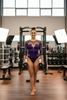 Female gymnast in purple leotard standing in a workout gym holding weights in her hands.