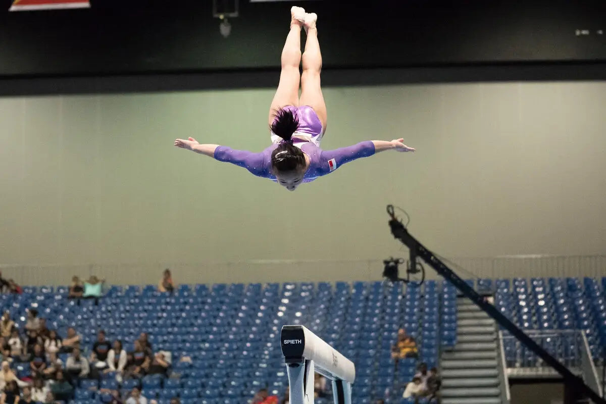A purple and white gymnast leotard with a small flag emblem on the back worn mid-air at artistic gymnastics championships